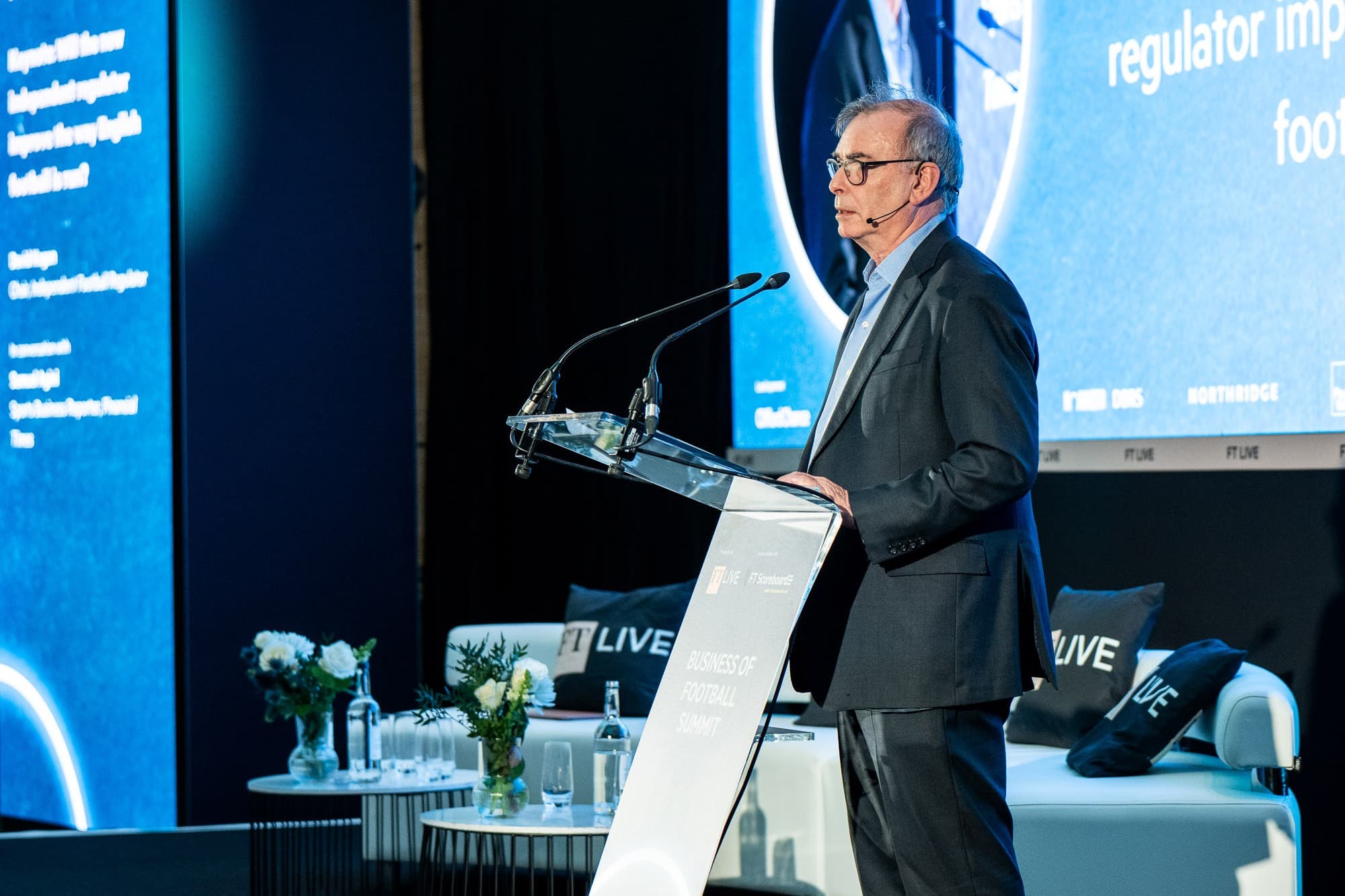 David Kogan speaking at the Financial Times Business of Football Summit. Stood at a glass lectern against a blue backdrop.
